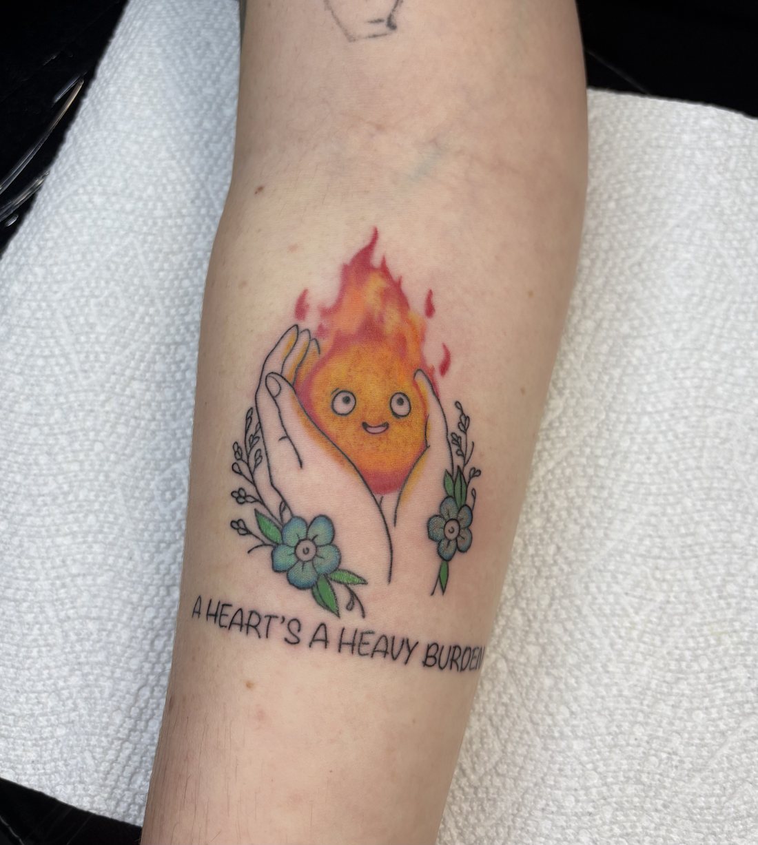 Fine-line color tattoo, a heart-shaped flame character held in two hands, with flowers, forearm